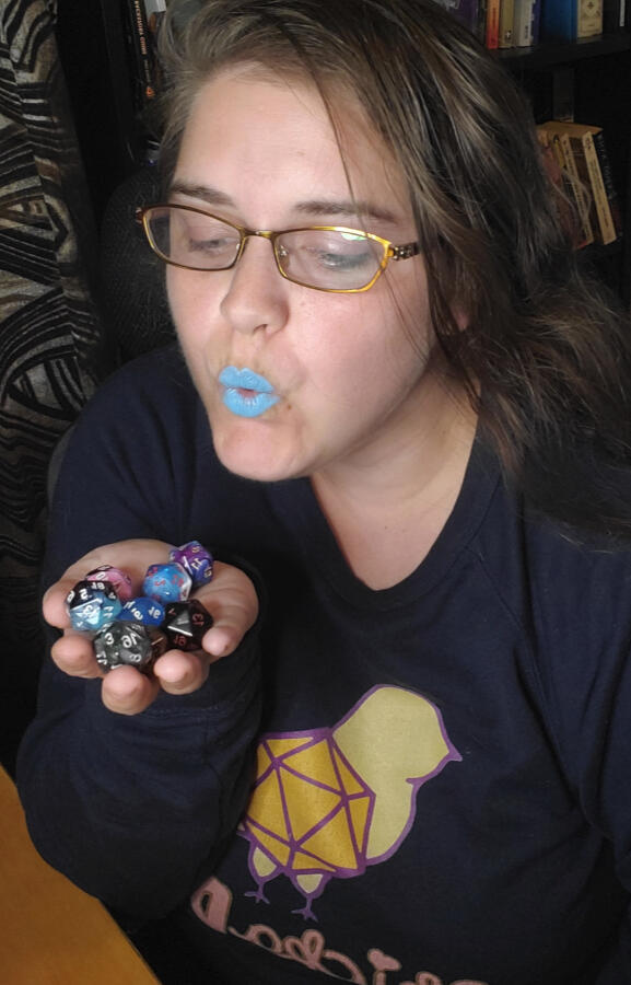 Chickadee, a white woman with brown hair wearing glasses and blue lipstick. Her lips are pursed, blowing on her dice filled hand. They are wearing a navy sweater with the ChickaDND logo.
